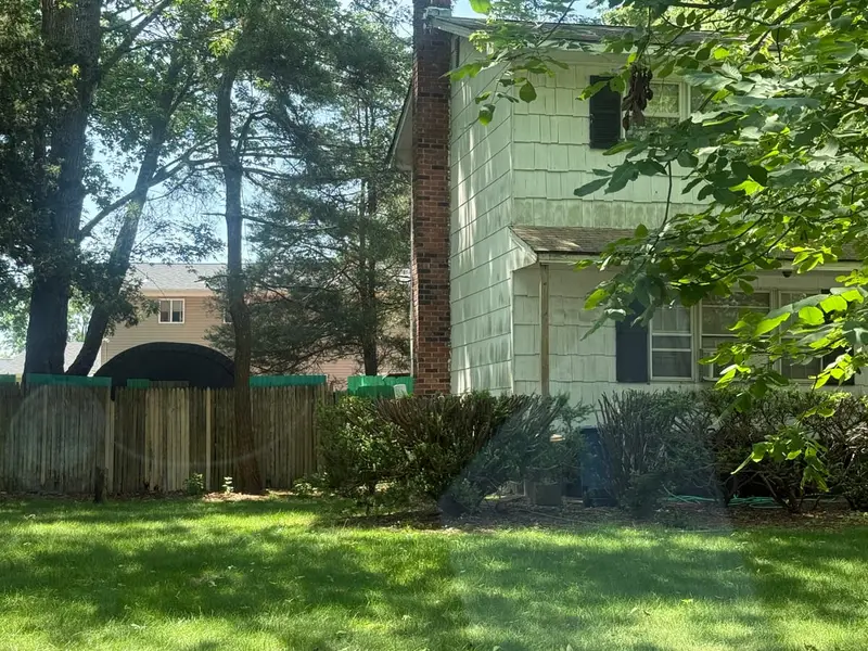 Two-story house with dark shutters behind a wood fence and shade trees, Patchogue, NY